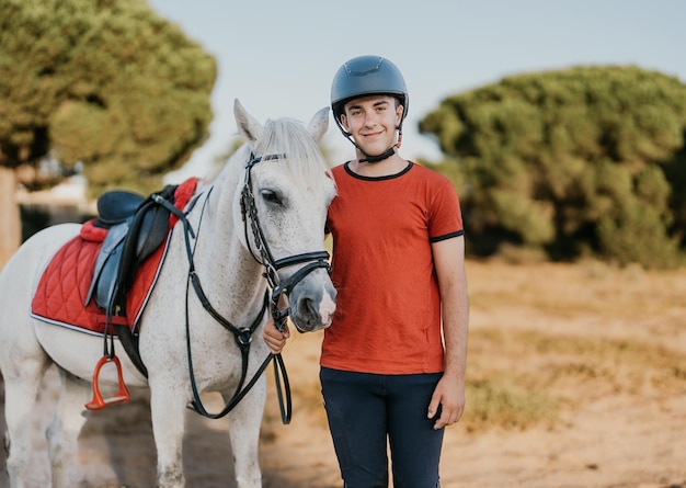 Portret van een jonge glimlachende ruiter die naast zijn witte paard in een dennenbos staat