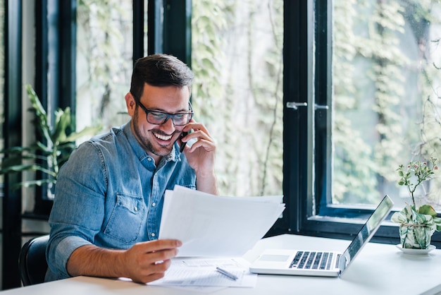 Portret van een jonge glimlachende, vrolijke ondernemer in een informeel kantoor die telefoongesprekken voert terwijl hij werkt met grafieken en grafieken die papieren documenten bekijken