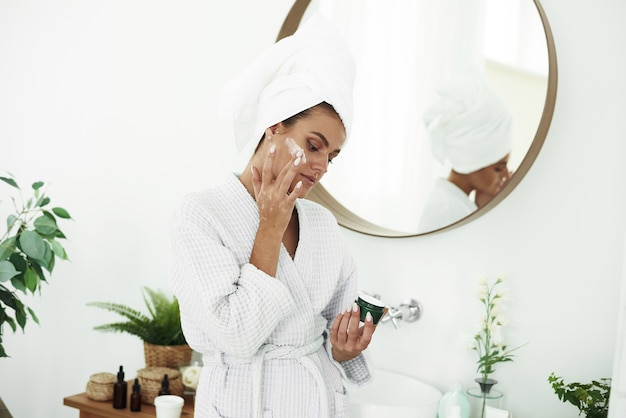 Portret van een jonge glimlachende vrouw die vochtinbrengende crème op haar gezicht in de badkamers toepast. Cosmetologie. Schoonheid en kuuroord.