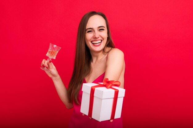 Portret van een jonge glimlachende vrouw in een roze stijlvolle jurk met een glas champagne en een kerstfeestje in een geschenkdoos