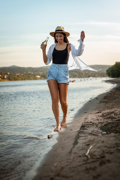 Portret van een jonge glimlachende vrouw in strohoed die bij zonsondergang op de rivierbank ontspant. Ze loopt langs het rivierstrand en lacht.