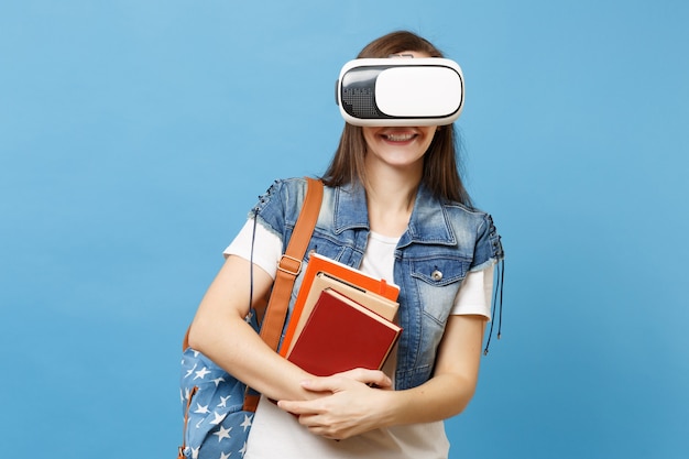 Portret van een jonge glimlachende vrouw student in denim kleding met rugzak met een virtual reality-bril, met schoolboeken geïsoleerd op een blauwe achtergrond. Onderwijs in middelbare school hogeschool.