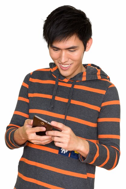 Portret van een jonge knappe Aziatische man glimlachend en met behulp van mobiele telefoon geïsoleerd tegen een witte muur