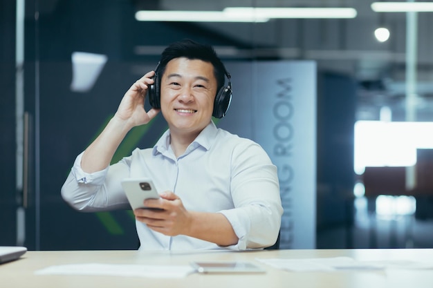 Portret van een jonge knappe aziatische man op kantoor, hij zit aan zijn bureau met een koptelefoon op