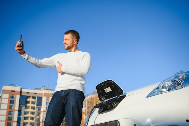 Portret van een jonge knappe europese man, die een selfie-foto maakt op zijn smartphone terwijl hij op zijn elektrische auto leunt, de batterij oplaadt bij de elektriciteitscentrale van de stad na het kopen van verse producten in het handelscentrum