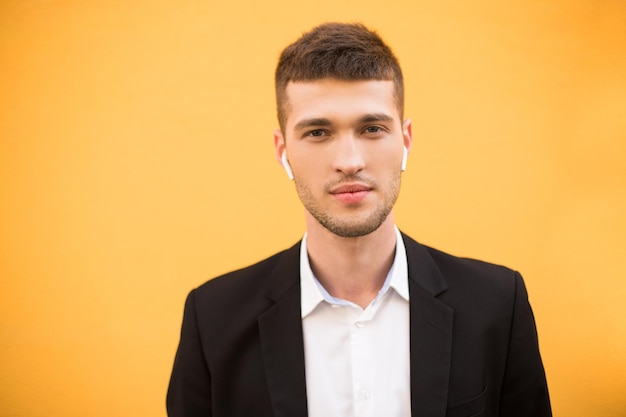 Portret van een jonge knappe man in klassieke zwarte jas en wit overhemd met draadloze koptelefoon die zorgvuldig in de camera kijkt over oranje achtergrond