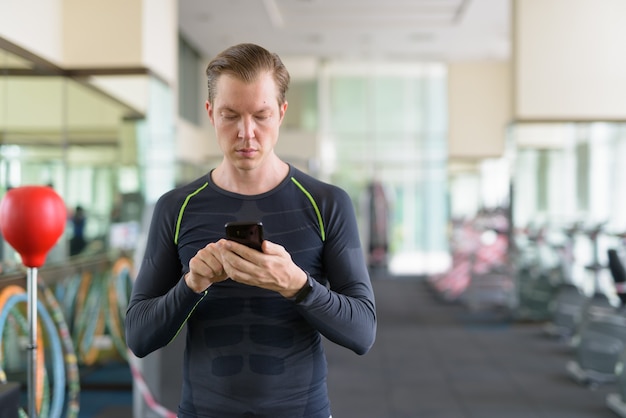 Portret van een jonge knappe man met behulp van telefoon in de sportschool tijdens covid-19