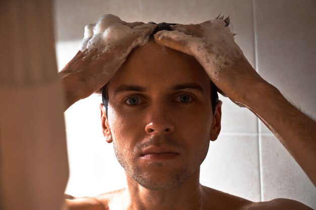 Portret van een jonge knappe man wast zichzelf met douchegel, schuimt het hoofd met shampoo in de badkamer thuis close-up