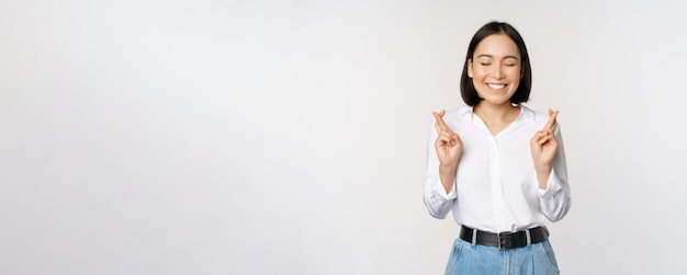 Portret van een jonge Koreaanse vrouw, een aziatisch meisje dat de vingers kruist en bidt en een wens maakt, anticiperend op resultaten die op een witte achtergrond staan