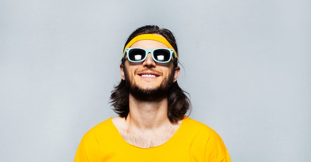 Portret van een jonge lachende man met lang haar met een zonnebril, een geel shirt en een band op het hoofd. Achtergrond van grijze getextureerde muur.