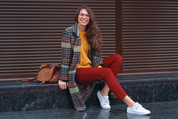 Portret van een jonge, lachende vrouw, gekleed in een stijlvolle jas, rode broek, oranje trui, witte sneakers, met een bruine leren tas, poseren in de straat van een Europese stad. Kopieer ruimte