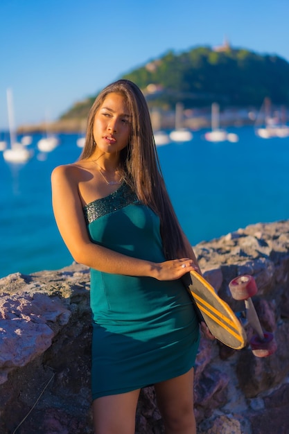 Portret van een jonge Latijnse brunette vrouw met een skate in een groene jurk met de zee op de achtergrond