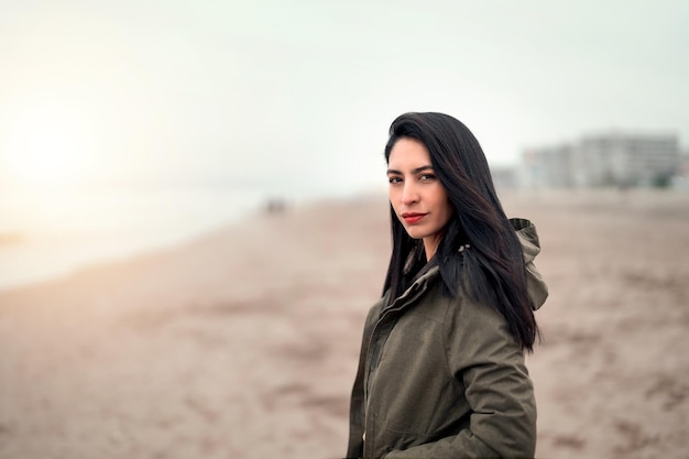 Portret van een jonge latijnse vrouw met een jas op het strand die naar de camera kijkt