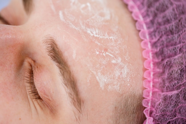 Portret van een jonge man bij het reinigen van de gezichtsprocedure door een schoonheidsspecialist van een vrouw in een schoonheidskliniek. De schoonheidsspecialist veegt het gezicht van de patiënt af met wattenschijfjes. Schoonheidsindustrie concept. Guy ligt op de bank.