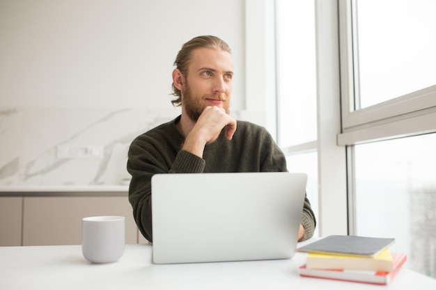 Portret van een jonge man die aan de tafel zit met een kopje en een laptop terwijl hij dromerig naar het raam kijkt