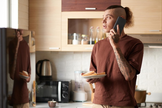 Portret van een jonge man die aan de telefoon praat terwijl hij in de keuken staat en een bord met broodjes vasthoudt
