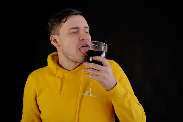 Portret van een jonge man die glas likt met rode wijn op zwarte achtergrond Gepassioneerde man in gele hoodie met wijnglas op donkere achtergrond