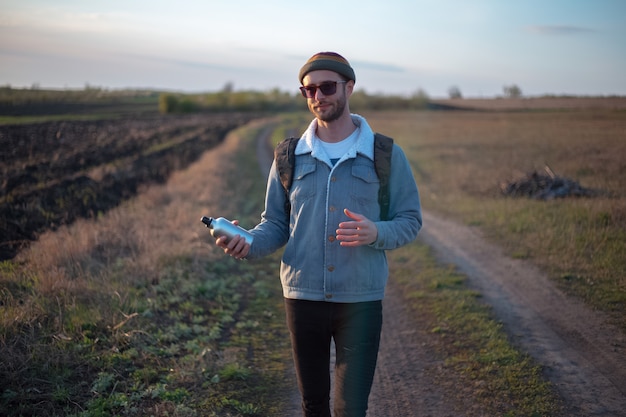 Portret van een jonge man die met rugzak herbruikbare aluminium thermo-waterfles in de hand houdt