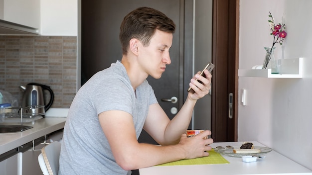 Portret van een jonge man heeft een ontbijt. Hij drinkt een kopje thee en bladert door de telefoon terwijl hij in een kleine keuken zit.