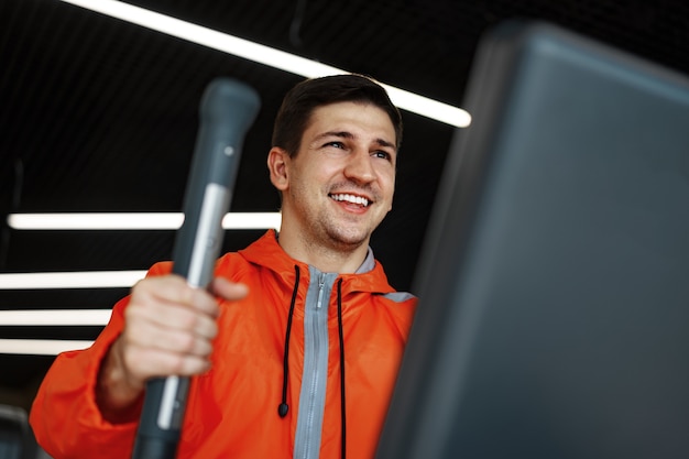 Portret van een jonge man in een oranje windjack die traint op een fitnessapparaat in de sportschool