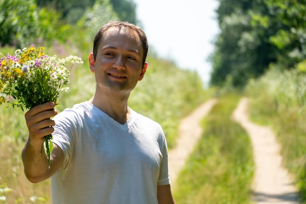 Portret van een jonge man in een wit T-shirt en met een boeket bloemen in zijn handen Een man poseert tegen de achtergrond van een bloemenveld en een onverharde weg in het dorp Het gezicht van een gelukkig persoon