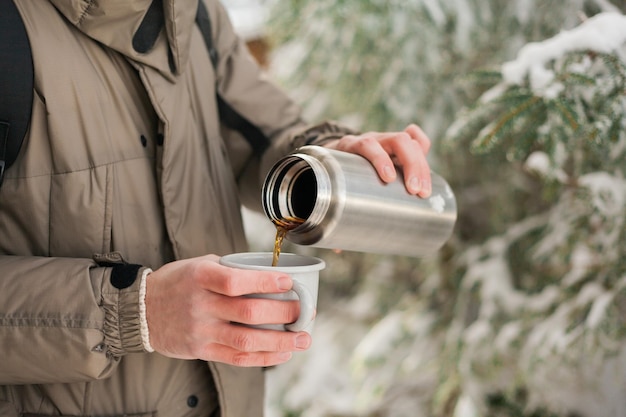 Portret van een jonge man in warme kleren in het winterbos Buiten hete thee drinken uit thermoskan Wandeltoerisme