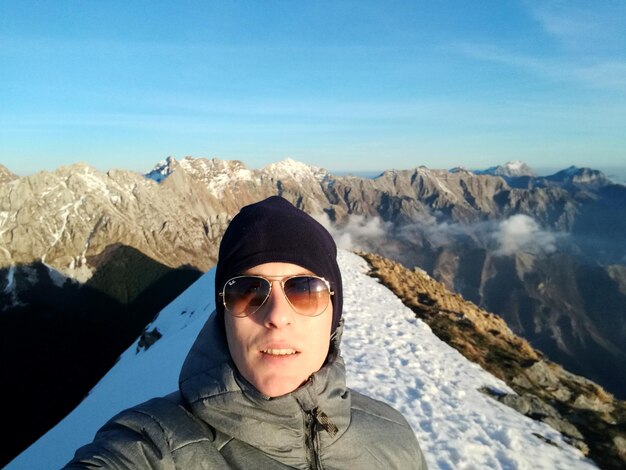 Foto portret van een jonge man met een zonnebril die in de winter tegen een bergketen staat