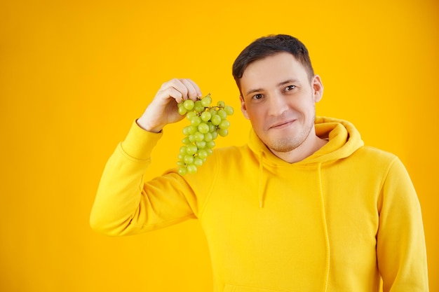 Portret van een jonge man met groene druiven op gele achtergrond Close-up van een knappe man in gele hoodie houdt een tros druiven vast