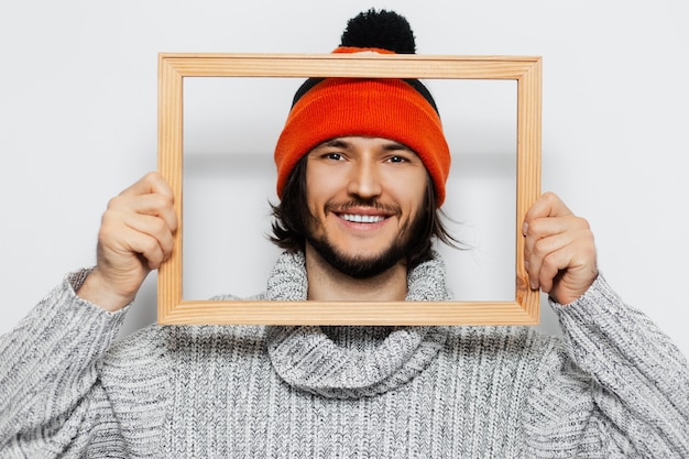 Portret van een jonge man met houten frame op zijn gezicht, oranje hoed en coltrui dragen op witte muur.