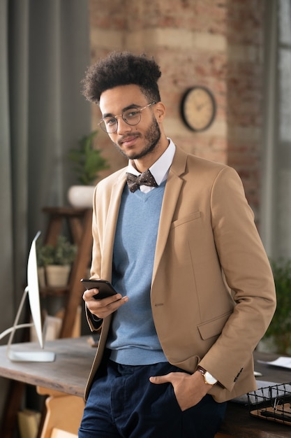 Portret van een jonge man met stijlvol kapsel die naar de camera kijkt terwijl hij zijn mobiele telefoon gebruikt