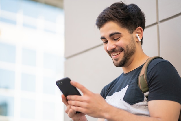 Portret van een jonge man met zijn mobiele telefoon terwijl hij buiten op straat staat.