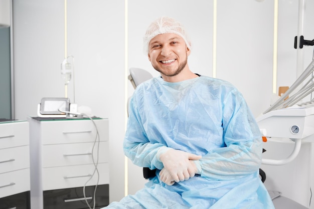 Portret van een jonge mannelijke tandarts die wegwerpkleding draagt en in een moderne kliniek zit
