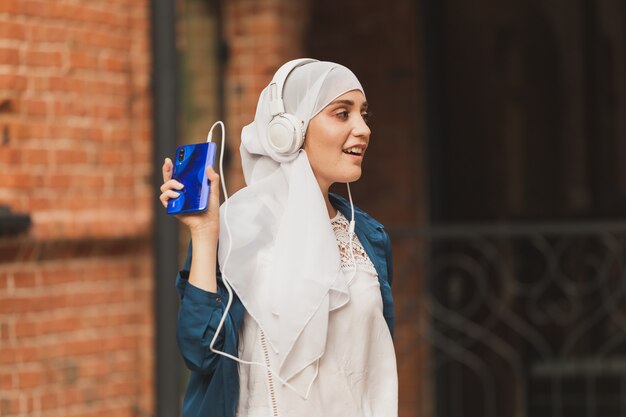 Portret van een jonge moderne arabische vrouw die een mobiele telefoon vasthoudt en de muziek luistert naar een hoofdtelefoon.