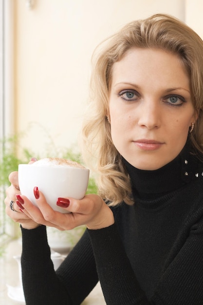 Portret van een jonge mooie blonde met blauwe ogen die koffie drinkt en rouwt bij het raam