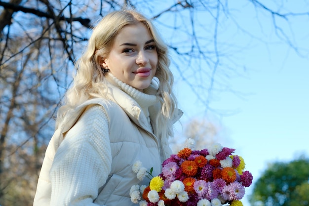 Portret van een jonge mooie blonde met lang haar in een witte trui en jas in een zonnige herfst in een stadspark met een boeket bloemen