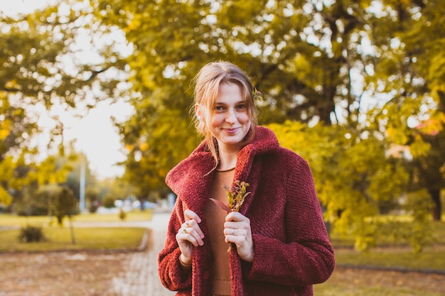 Portret van een jonge, mooie glimlachende vrouw die in het herfstpark staat met revers van haar donkerrode jas. Mooi meisje met kleine bloemen in haar haar, glimlachend, kijkend naar de camera.