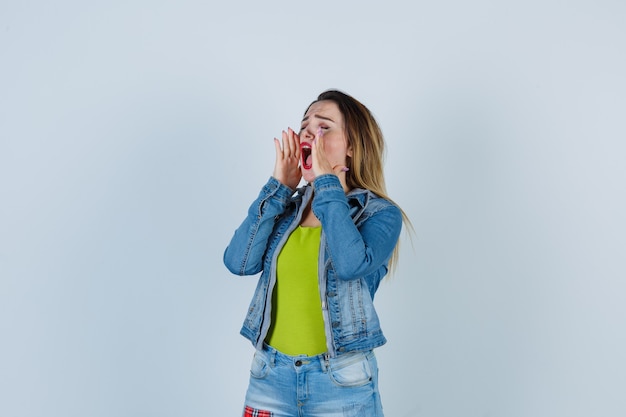 Portret van een jonge, mooie vrouw die de handen in de buurt van de mond houdt in een denim-outfit en er een verontrust vooraanzicht uitziet