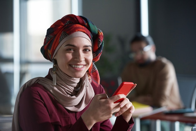 Portret van een jonge moslimzakenvrouw die naar de camera glimlacht terwijl ze een bericht leest op haar mobiele telefoon op kantoor