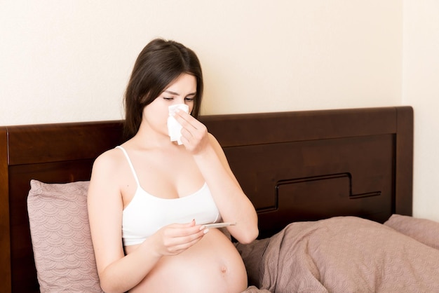 Portret van een jonge ongelukkige zwangere vrouw die op kussens op bed leunt en haar neus in het weefsel snuit. Toekomstige moeder werd verkouden en niesde