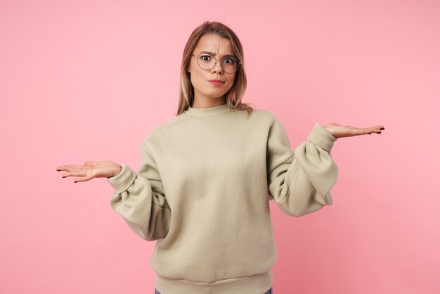 Portret van een jonge ontevreden vrouw in een bril die zich voordeed met het overgeven van handen geïsoleerd op roze