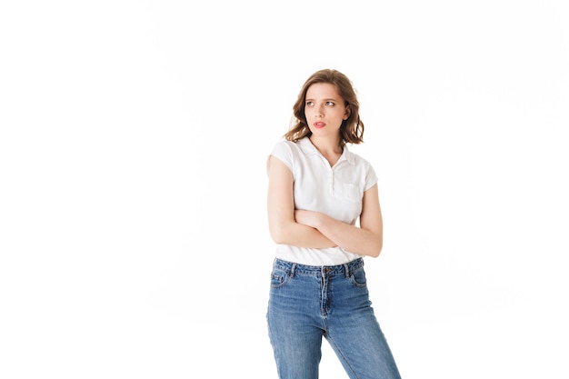 Portret van een jonge overstuur dame die in een t-shirt en spijkerbroek staat en helaas opzij kijkt op een witte achtergrond geïsoleerd