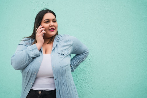 Portret van een jonge plus size vrouw die lacht terwijl ze buiten telefoneert tegen een lichtblauwe muur. Communicatieconcept.