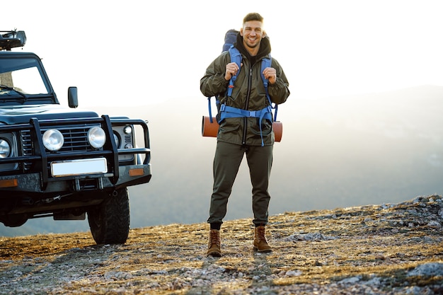 Portret van een jonge reizigersmens in wandeluitrusting die zich dichtbij zijn off-road auto bevindt