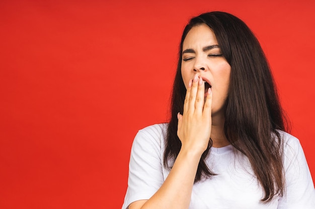 Portret van een jonge, schattige, vermoeide vrouw die geeuwt met haar ogen dicht en haar mond bedekt, geïsoleerd over rode achtergrond