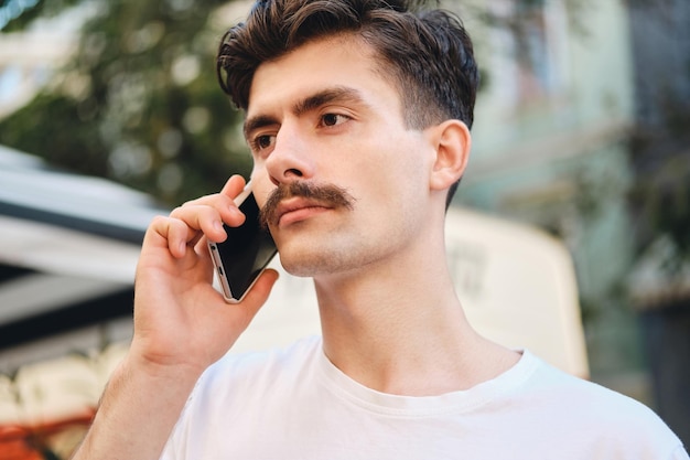 Portret van een jonge serieuze snorman in een T-shirt die op zijn mobiel praat terwijl hij emotieloos opzij kijkt op straat in de stad