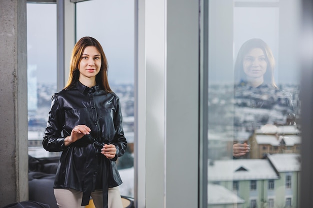 Portret van een jonge slanke vrouw in een bril en een zwart leren overhemd Moderne vrouw op de achtergrond van het raam op kantoor met een groot raam