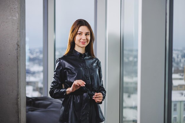 Portret van een jonge slanke vrouw in een bril en een zwart leren overhemd Moderne vrouw op de achtergrond van het raam op kantoor met een groot raam