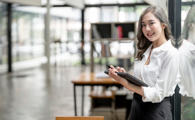 Portret van een jonge smiley-aziatische zakenvrouw die een tablet vasthoudt in een moderne officier die glimlacht en naar de camera kijkt