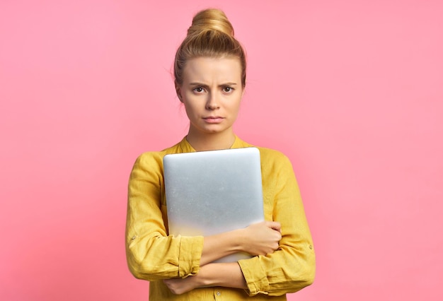 Portret van een jonge student die een laptop in haar armen houdt terwijl ze lacht geïsoleerd tegen een roze achtergrond