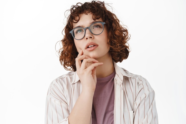 Portret van een jonge studente die denkt, een vrouw met een bril die nadenkend opkijkt, nadenkt, een raadsel oplost, naar de bovenste advertentie staart en een keuze maakt, witte achtergrond